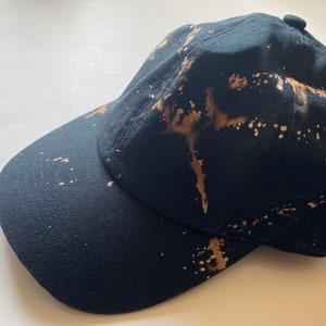 Könnte beinhalten: Eine schwarze Baseballkappe mit einem bleichmittelgesprenkelten Design in Braun- und Weißtönen.