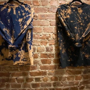 Könnte beinhalten: Zwei langärmelige Hemden mit einem Batik-Muster. Ein Hemd ist marineblau mit beigefarbenen Bleichflecken, das andere ist schwarz mit beigefarbenen Bleichflecken.