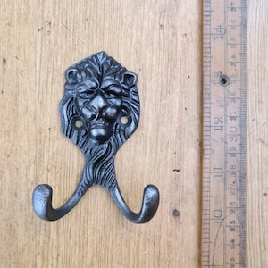 May include: A cast iron double hook with a lion's head design. The dark grey hook has two curved hooks below the lion's head. The lion's face has detailed features. The hook is approximately 13 cm long.