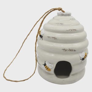 May include: Cream ceramic beehive-shaped birdhouse with a brown rope. The birdhouse has a round entrance and is decorated with three cartoon bees with yellow and black stripes. A decorative garden ornament.