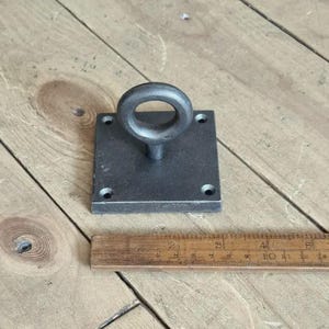 May include: A dark grey metal ring pull with a circular handle and a square base with four mounting holes. A wooden ruler is next to the ring pull. The ring pull is likely for a door or drawer.