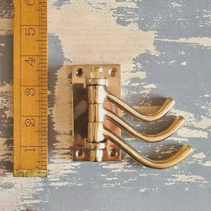 May include: A brass triple coat hook on a rectangular plate. The curved hooks extend outwards. A wooden ruler is next to the hook, showing measurements in inches. The hook is approximately 10 cm in length.