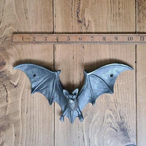 May include: A cast metal bat wall decoration in a dark grey finish. The bat has outstretched wings and is approximately 28 cm wide. The bat is mounted on a wooden surface with a ruler.