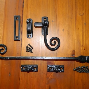 May include: Black wrought iron window or door latch hardware set. The set includes a long bar with a decorative scroll end, a catch, and mounting plates. The hardware is shown on a wooden surface, with screws included.