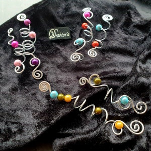 Könnte beinhalten: Drei silberne Haarschmuckstücke aus Draht mit bunten Perlen. Das erste Stück hat lila, rosa und blaue Perlen. Das zweite Stück hat türkisfarbene, rote und orange Perlen. Das dritte Stück hat türkisfarbene, grüne und gelbe Perlen.