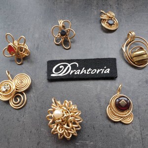 Könnte beinhalten: Eine Sammlung von sieben goldenen, drahtgewickelten Anhängern mit verschiedenen farbigen Steinen. Die Anhänger haben unterschiedliche Designs, darunter Blumen, Spiralen und Kreise. Das Wort "Drahtoria" ist auf einem schwarzen Etikett in der Mitte des Bildes aufgedruckt.
