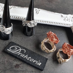 Könnte beinhalten: Drei Drahtringe, einer silber, einer gold und einer kupfer, auf schwarzen Ringkegeln ausgestellt. Die Ringe sind aus verdrehtem Draht gefertigt und haben ein Spiraldesign. Die Ringe sind in einer Reihe auf einer weißen Oberfläche mit einem schwarzen Etikett angeordnet, auf dem "Drahtoria" steht.