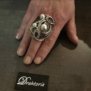 Könnte beinhalten: Ein großer Statement-Ring mit einem komplexen Design aus gewickeltem, silberfarbenem Draht und drei glatten, runden, grauen Perlen. Der Ring befindet sich in der Nähe eines schwarzen, rechteckigen Etiketts mit dem weißen Schriftzug Drahtoria.
