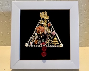 May include: A white framed picture with a black background featuring a Christmas tree made of various jewelry and trinkets. The tree is decorated with rhinestones, pearls, and other small ornaments.