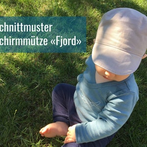 Può includere: Un bambino con un berretto grigio chiaro con visiera è seduto sull'erba verde. Il bambino indossa una maglietta blu a maniche lunghe e pantaloni blu scuro. Il testo su un rettangolo verde recita "Schnittmuster Schirmmütze «Fjord»".