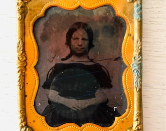 May include: Antique portrait photograph in a decorative gold-toned frame. The image features a woman in a dark dress with her hands clasped. The frame has an ornate design with an orange interior and gold exterior.