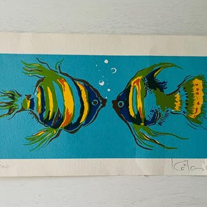 May include: A colorful print featuring two stylized fish facing each other against a turquoise background. The fish have yellow, green, and blue stripes and accents. Bubbles float above the fish. The print is numbered 142/200 and signed.