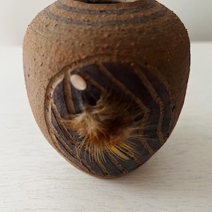 Puede incluir: Pequeño jarrón de cerámica artesanal con superficie marrón texturizada. Presenta una abertura tallada que revela un interior oscuro y un mechón de fibras marrones y amarillas. El diseño incluye bandas horizontales en la parte superior.
