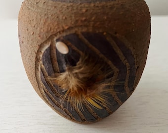 May include: A small, handcrafted ceramic vase with a textured, brown surface. The vase features a carved opening revealing a dark interior and a tuft of brown and yellow fibers. The design includes horizontal bands around the top.