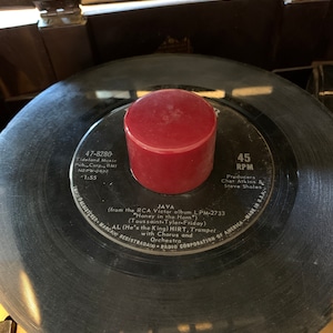 Puede incluir: Un disco de vinilo de 45 RPM vintage con un objeto cilíndrico rojo encima. La etiqueta del disco presenta el título de la canción "JAVA" e información del artista. El disco es negro con surcos visibles.