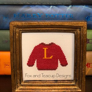 May include: A framed cross-stitch of a red sweater with a yellow letter L on it. The frame is gold and the cross-stitch is on white fabric. The frame is sitting on a stack of Harry Potter books.