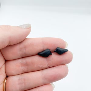 May include: Two small, black bird-shaped earrings. The earrings are held in a hand against a white background. The earrings are made of a smooth, glossy material.