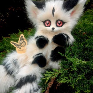 May include: A plush creature with long white and black faux fur, large pointed ears, and red eyes featuring black heart-shaped pupils. The creature has black markings on its forehead and paws, and a tan butterfly-shaped tag attached to its side.