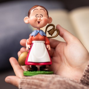 Puede incluir: Una figurita de una mujer con una cabeza grande y una gran sonrisa. Lleva un vestido rojo con un delantal blanco y una bufanda azul y blanca. Lleva un huevo dorado en su mano izquierda y una campana en su mano derecha.