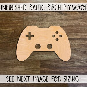 May include: Unfinished Baltic birch plywood cutout of a video game controller. The controller is light brown and has a plus sign, four circles, and a circle with a cross in the center.