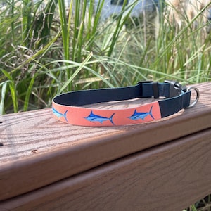 Könnte beinhalten: Ein dunkelblaues Haustierhalsband mit einem korallenfarbenen Abschnitt, der mit einem sich wiederholenden Muster aus blauen Marlinfischen verziert ist. Das Halsband verfügt über eine schwarze Kunststoff-Steckschnalle und einen silbernen D-Ring.