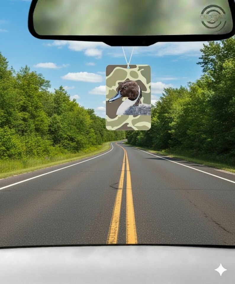 Pode incluir: Ambientador retangular com uma ilustra&ccedil;&atilde;o de pato sobre um padr&atilde;o de camuflagem. O ambientador est&aacute; pendurado num espelho retrovisor, com uma estrada e &aacute;rvores ao fundo. O pato tem a cabe&ccedil;a castanha e o bico azul.