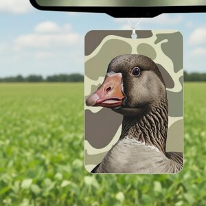 May include: Rectangular air freshener featuring a detailed illustration of a goose's head and neck, set against a camouflage pattern. The goose has a brown head, a pinkish-orange beak, and a gray neck. The air freshener hangs from a string.