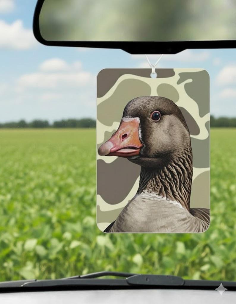May include: Rectangular air freshener featuring a detailed illustration of a goose's head and neck, set against a camouflage pattern. The goose has a brown head, a pinkish-orange beak, and a gray neck. The air freshener hangs from a string.