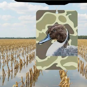 May include: Rectangular air freshener featuring a detailed illustration of a duck with a brown head, blue beak, and white chest, set against a camouflage background. The air freshener is hanging in front of a field of harvested crops.