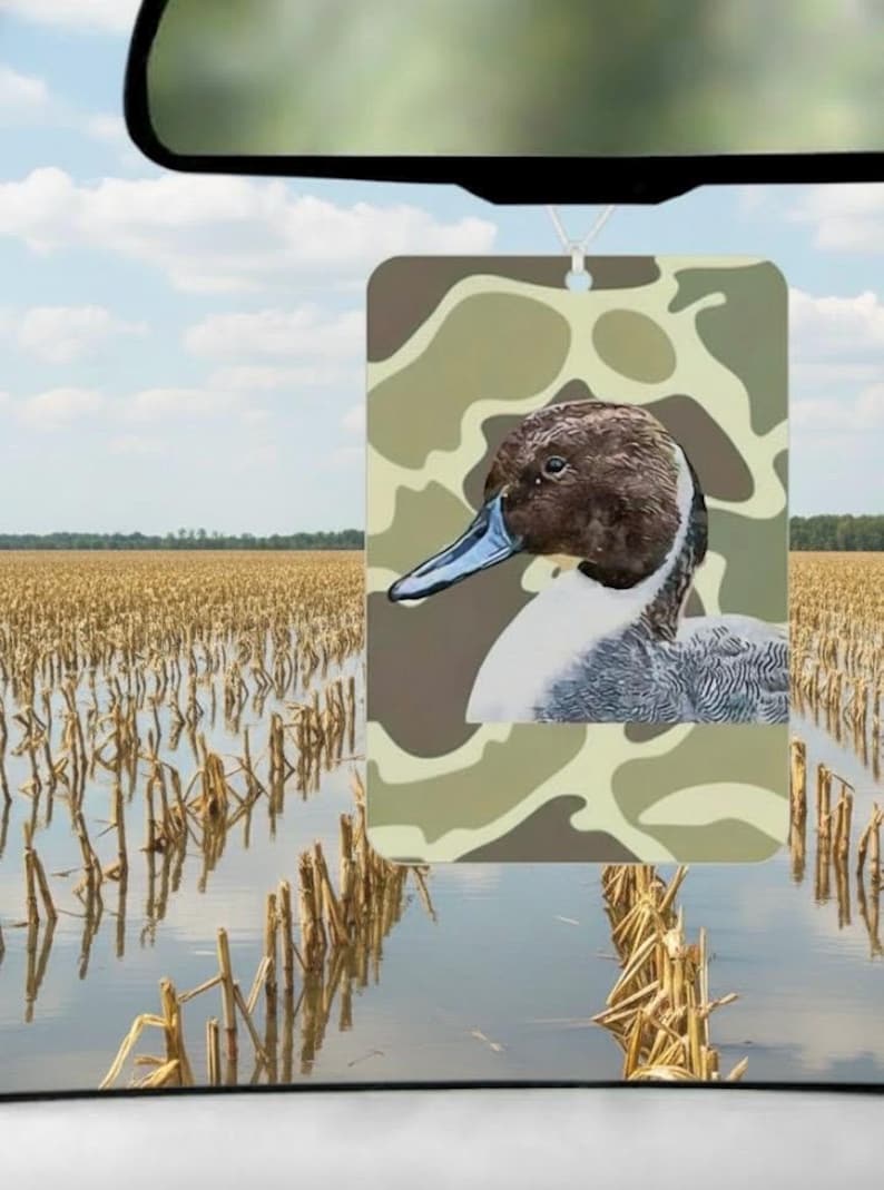 May include: Rectangular air freshener featuring a detailed illustration of a duck with a brown head, blue beak, and white chest, set against a camouflage background. The air freshener is hanging in front of a field of harvested crops.