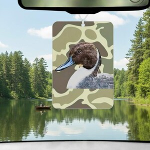 Pode incluir: Ambientador retangular com uma ilustra&ccedil;&atilde;o de pato, cabe&ccedil;a castanha e bico azul, sobre um fundo de camuflagem. Pendurado no espelho retrovisor de um carro, com uma cena de lago ao fundo. Projetado para refrescar o ar dentro de um ve&iacute;culo.