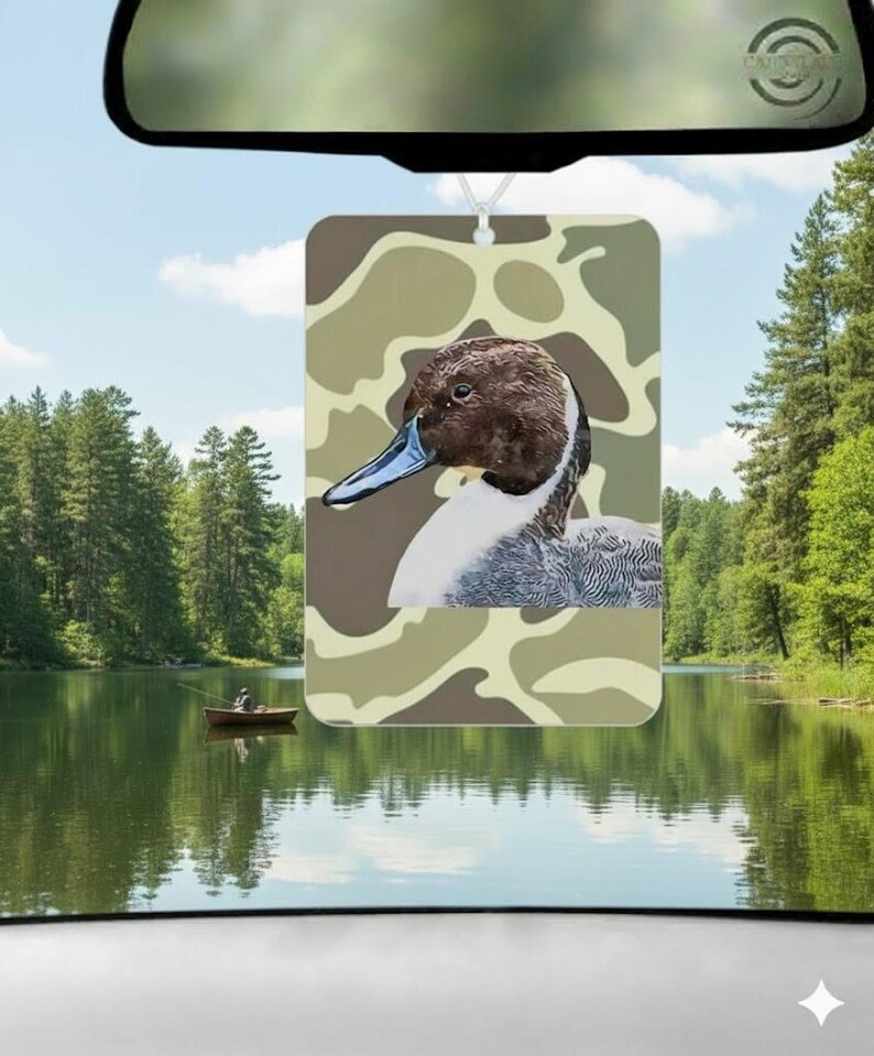 Pode incluir: Ambientador retangular com uma ilustra&ccedil;&atilde;o de pato, cabe&ccedil;a castanha e bico azul, sobre um fundo de camuflagem. Pendurado no espelho retrovisor de um carro, com uma cena de lago ao fundo. Projetado para refrescar o ar dentro de um ve&iacute;culo.