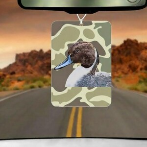May include: Rectangular car air freshener featuring a duck illustration with a camouflage background. The duck has a brown head, blue beak, and white and gray feathers. The air freshener hangs from a rearview mirror against a scenic road backdrop.