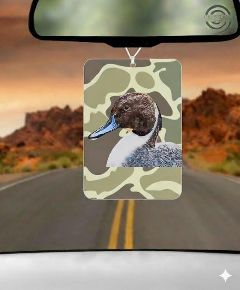 May include: Rectangular car air freshener featuring a duck illustration with a camouflage background. The duck has a brown head, blue beak, and white and gray feathers. The air freshener hangs from a rearview mirror against a scenic road backdrop.