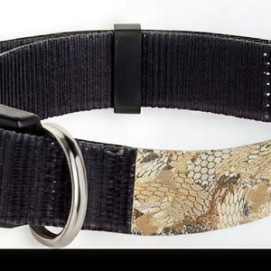 May include: Black dog collar with a camouflage pattern in tan and brown. The collar features a black plastic buckle and a silver metal D-ring for leash attachment. The collar is made of durable nylon material.