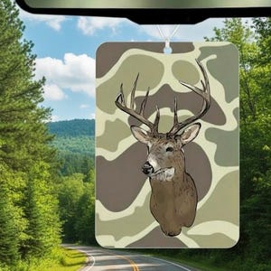 May include: Rectangular air freshener with a deer head illustration on a camouflage background in brown and green tones. The air freshener hangs from a car's rearview mirror, with a road and forest visible in the background.