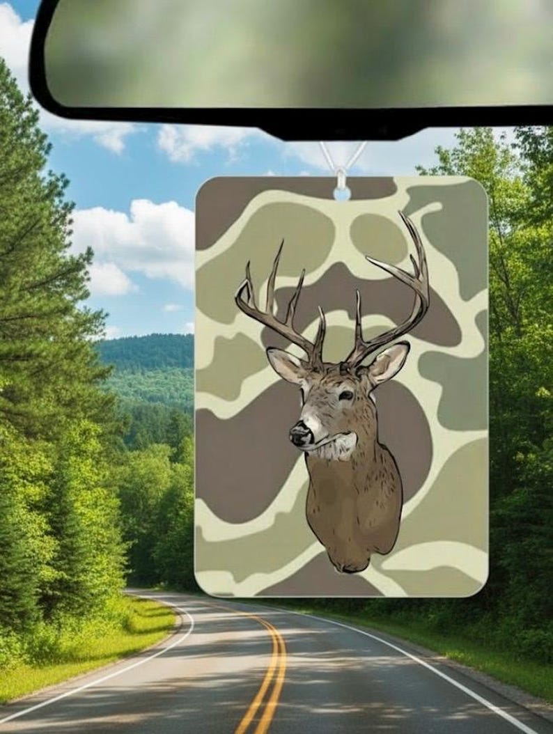 May include: Rectangular air freshener with a deer head illustration on a camouflage background in brown and green tones. The air freshener hangs from a car's rearview mirror, with a road and forest visible in the background.