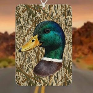 May include: Rectangular air freshener featuring a detailed illustration of a mallard duck's head. The duck has a green head, yellow beak, and brown neck. The background is a camouflage pattern of tall grass. A white string is attached for hanging.