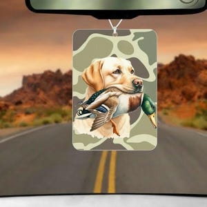 May include: Rectangular air freshener with a yellow Labrador Retriever holding a duck, set against a camouflage pattern. The image hangs from a string, with a road and mountain backdrop visible.