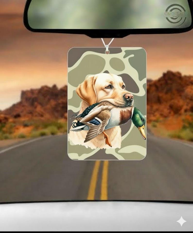 May include: Rectangular air freshener with a yellow Labrador Retriever holding a duck, set against a camouflage pattern. The image hangs from a string, with a road and mountain backdrop visible.