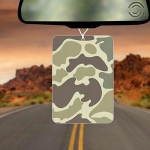 May include: Rectangular car air freshener with a camouflage pattern in shades of brown, tan, and green, hanging from a rearview mirror. The background shows a road leading towards a desert landscape.