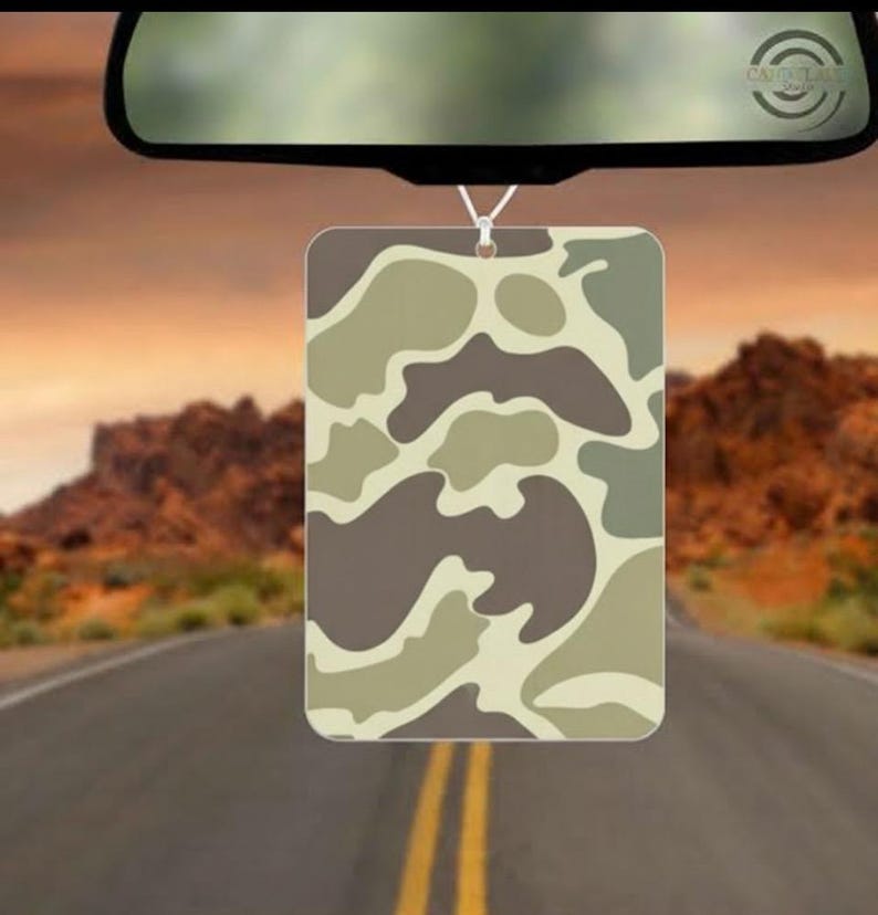 May include: Rectangular car air freshener with a camouflage pattern in shades of brown, tan, and green, hanging from a rearview mirror. The background shows a road leading towards a desert landscape.