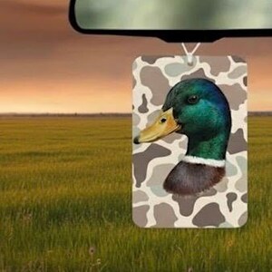 Pode incluir: Ambientador retangular com uma ilustra&ccedil;&atilde;o detalhada da cabe&ccedil;a de um pato-real. Possui penas verdes e castanhas iridescentes, um bico amarelo e um colar branco. O fundo apresenta um padr&atilde;o de camuflagem em tons de castanho e bege. O ambientador est&aacute; pendurado em frente a um campo.