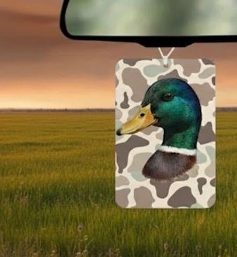 Pode incluir: Ambientador retangular com uma ilustra&ccedil;&atilde;o detalhada da cabe&ccedil;a de um pato-real. Possui penas verdes e castanhas iridescentes, um bico amarelo e um colar branco. O fundo apresenta um padr&atilde;o de camuflagem em tons de castanho e bege. O ambientador est&aacute; pendurado em frente a um campo.