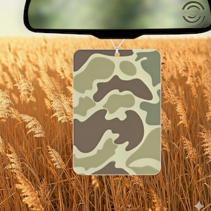 May include: Rectangular air freshener with a camouflage pattern in shades of brown, tan, and green, hanging from a rearview mirror. The background features tall, golden-brown grass.
