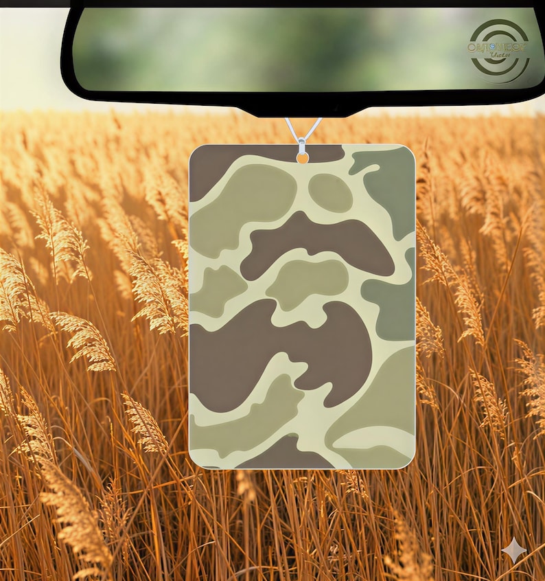 May include: Rectangular air freshener with a camouflage pattern in shades of brown, tan, and green, hanging from a rearview mirror. The background features tall, golden-brown grass.