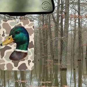 May include: A car air freshener featuring a mallard duck head with a green, blue, and brown color scheme. The duck is set against a camouflage background. The air freshener hangs from a car's rearview mirror, with a backdrop of a flooded forest.