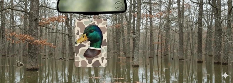 May include: A car air freshener featuring a mallard duck head with a green, blue, and brown color scheme. The duck is set against a camouflage background. The air freshener hangs from a car's rearview mirror, with a backdrop of a flooded forest.