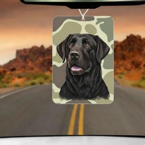 May include: Rectangular car air freshener featuring a black Labrador Retriever against a camouflage background. The air freshener is hanging from a rearview mirror, with a road and desert landscape visible in the background.