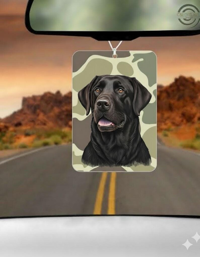 May include: Rectangular car air freshener featuring a black Labrador Retriever against a camouflage background. The air freshener is hanging from a rearview mirror, with a road and desert landscape visible in the background.